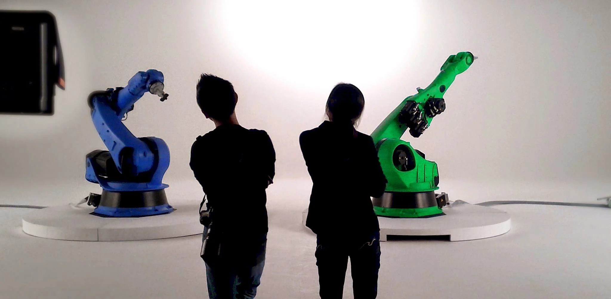 Zi and his wife, seen from behind, facing a blue KUKA robotic arm and a green KUKA robotic arm at ACME Lab.